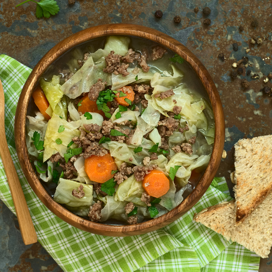 The Savoy Cabbage Soup in a wooden bowl on top of a light green plaid dish towel with toast and spoon on the side