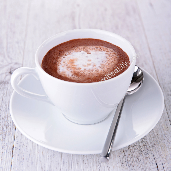 Hot chocolate with cream in white cup resting on a white saucer on a wood grain surface with silver spoon