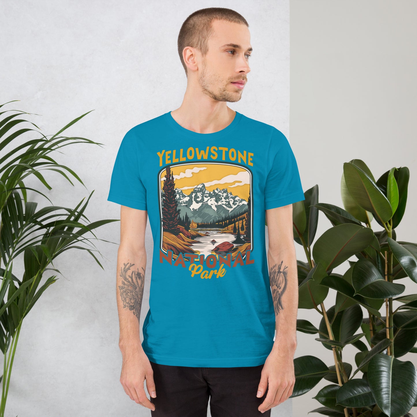 A male with head to the right wearing the Yellowstone National Park T-shirt in aqua against a wall with flowers