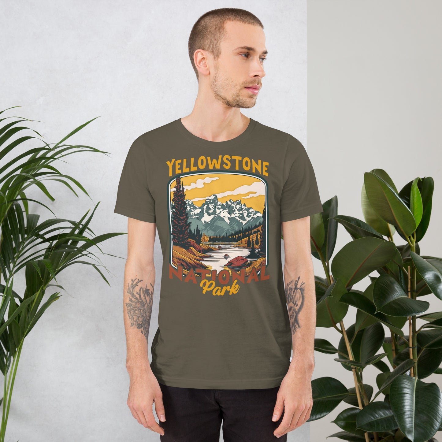 A male with head to the right wearing the Yellowstone National Park T-shirt in army green against a wall with flowers