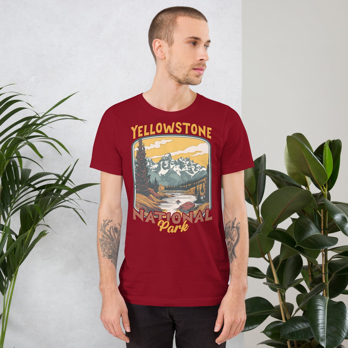 A male with head to the right wearing the Yellowstone National Park T-shirt in cardinal against a wall with flowers