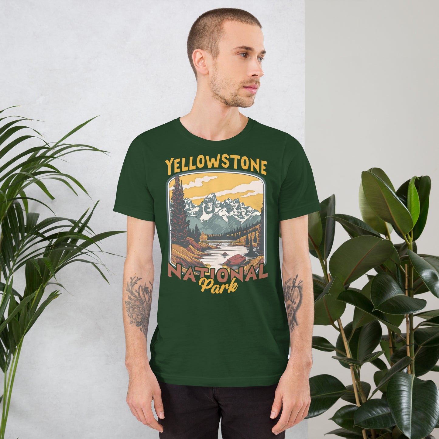 A male with head to the right wearing the Yellowstone National Park T-shirt in forest green against a wall with flowers