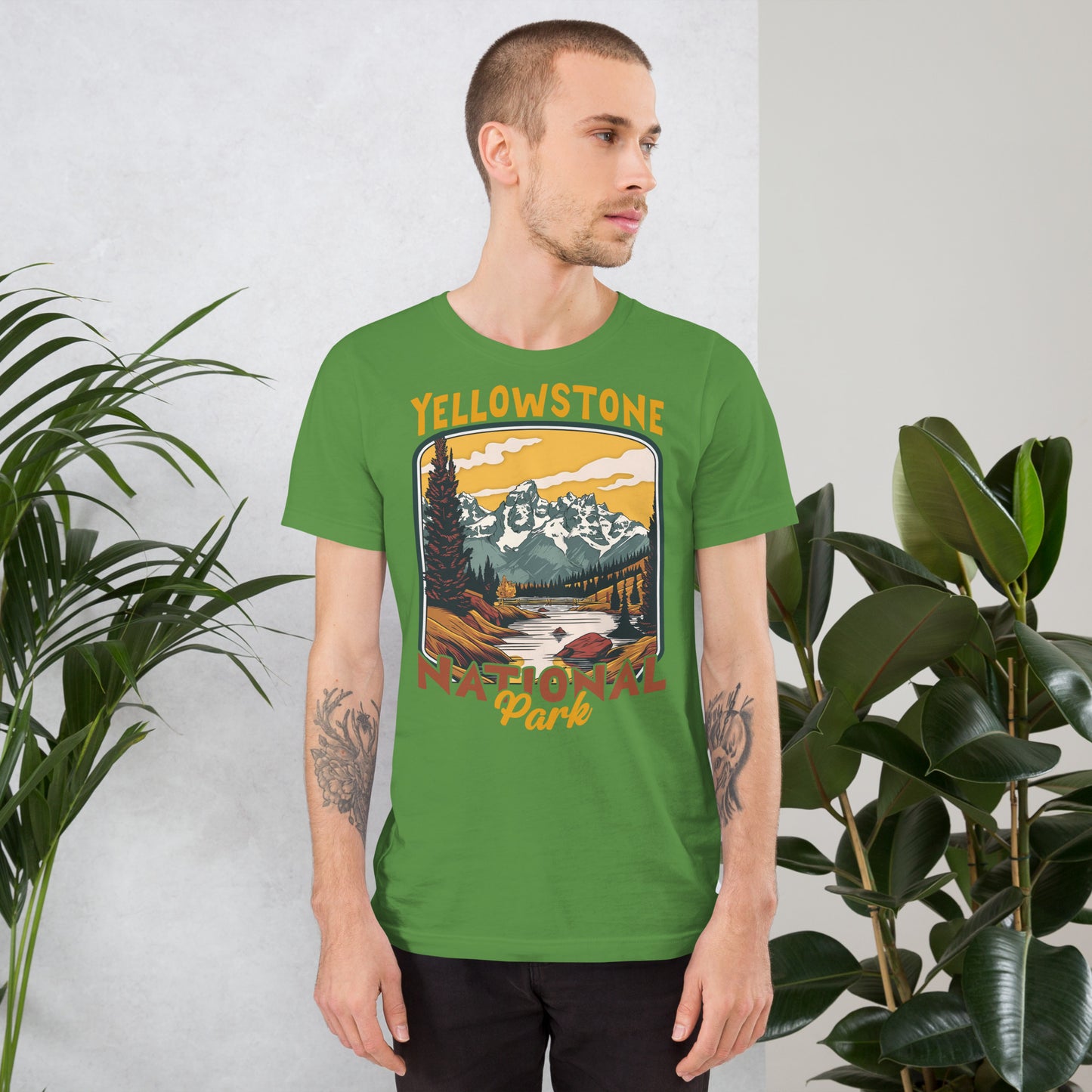 A male with head to the right wearing the Yellowstone National Park T-shirt in leaf green against a wall with flowers