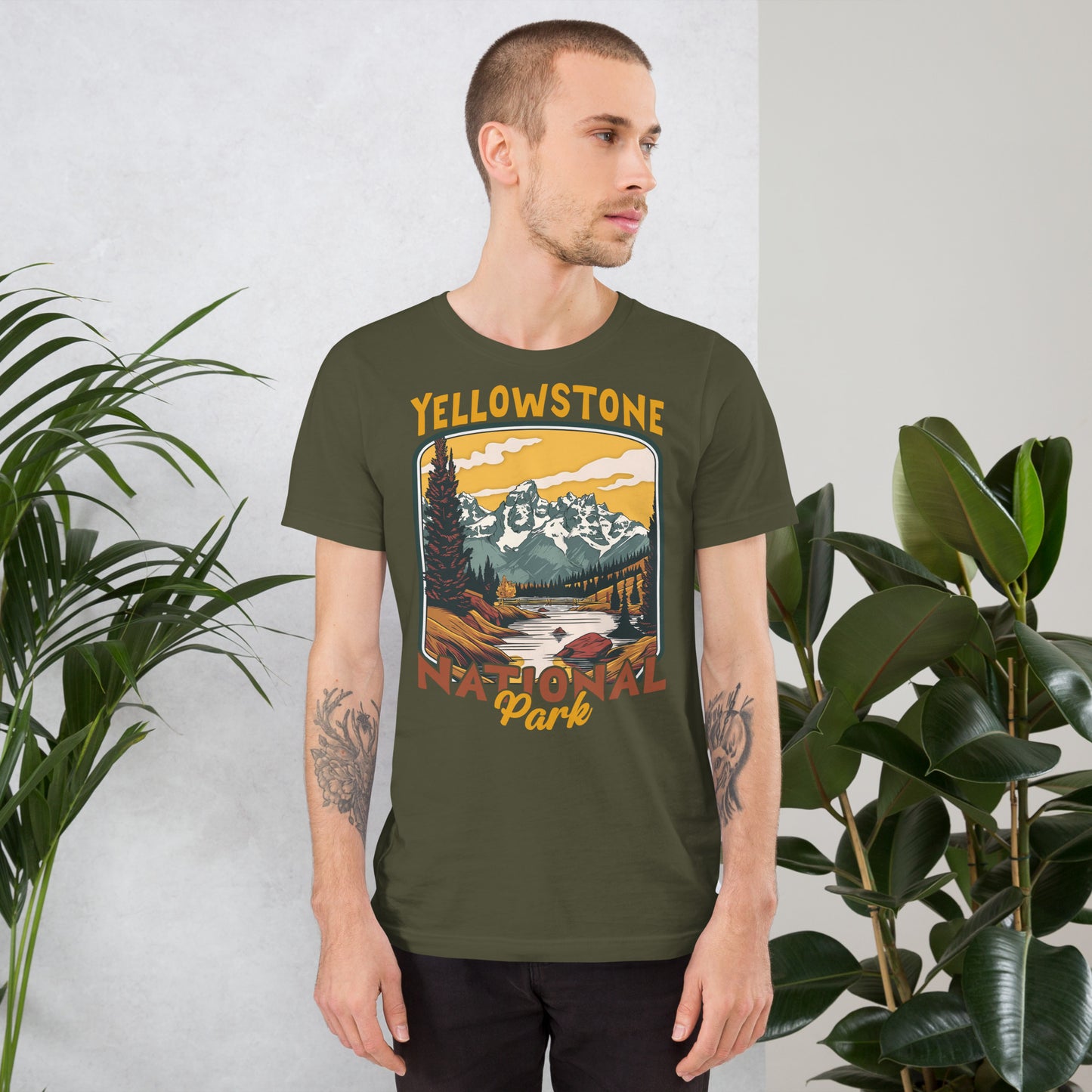 A male with head to the right wearing the Yellowstone National Park T-shirt in military green against a wall with flowers