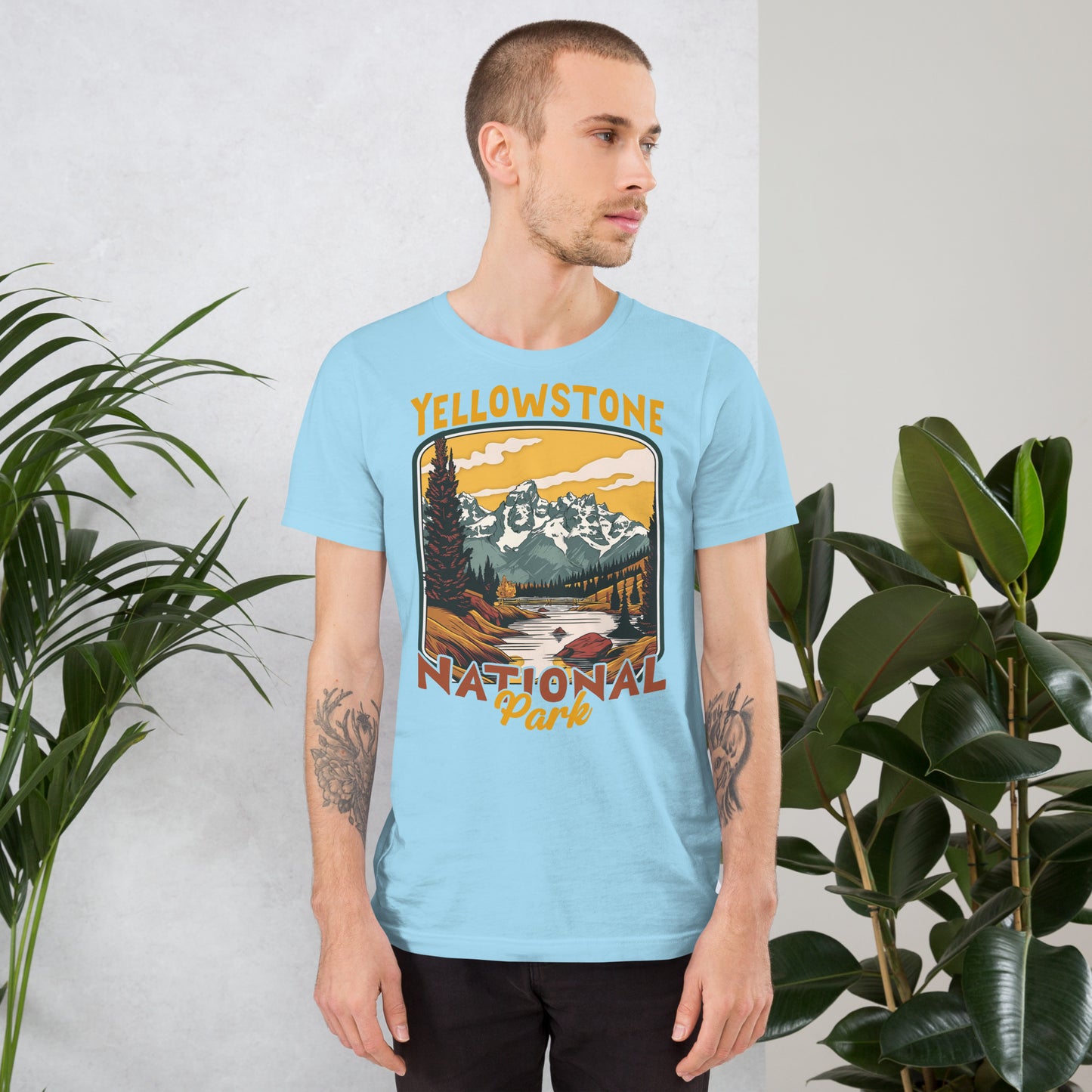 A male with head to the right wearing the Yellowstone National Park T-shirt in ocean blue against a wall with flowers