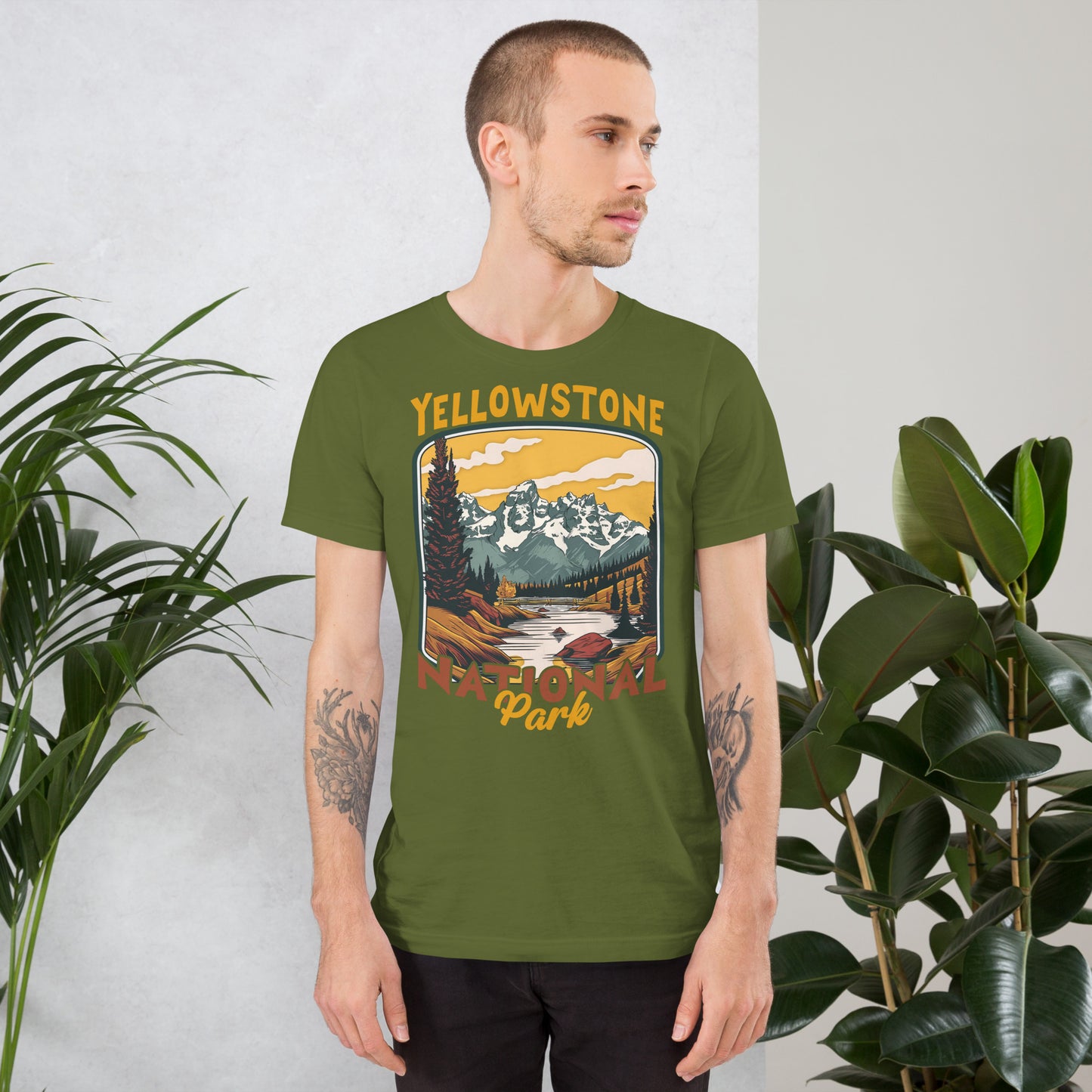 A male with head to the right wearing the Yellowstone National Park T-shirt in olive green against a wall with flowers