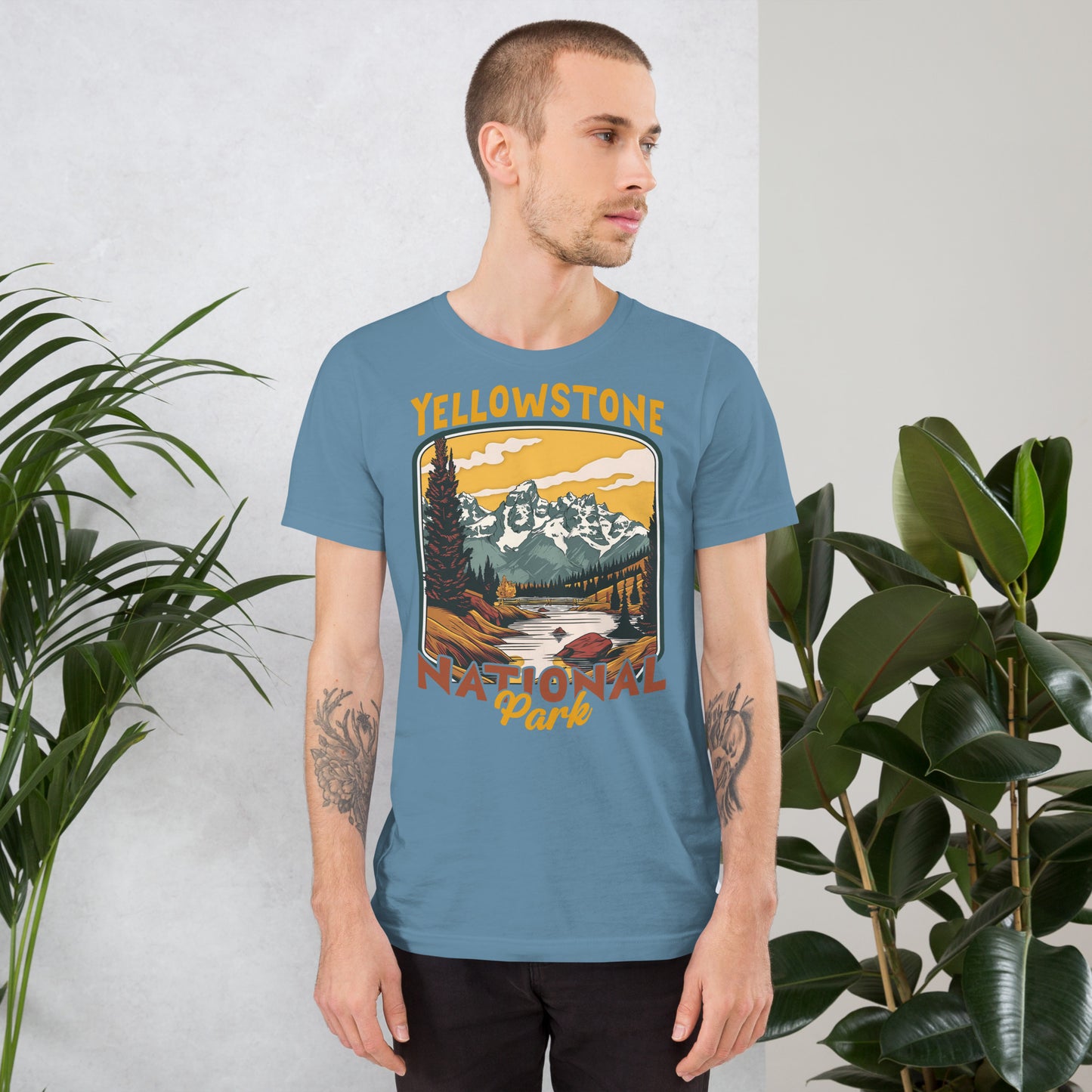 A male with head to the right wearing the Yellowstone National Park T-shirt in steel blue against a wall with flowers